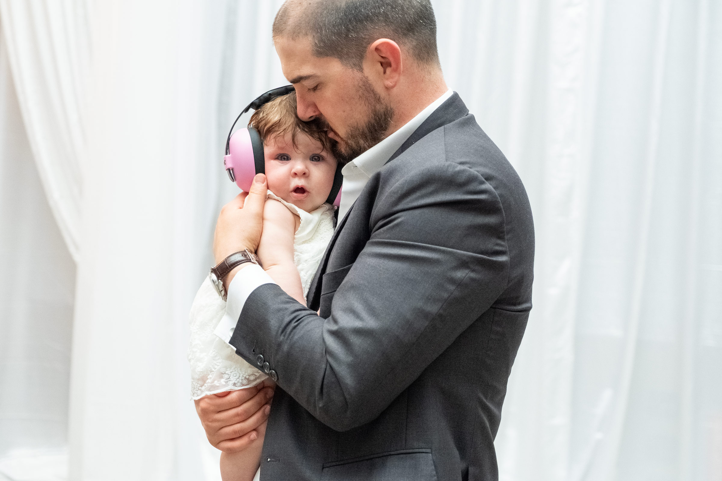 Groom in a grey suit gently holding his baby daughter, wearing pink ear protection, at a wedding reception