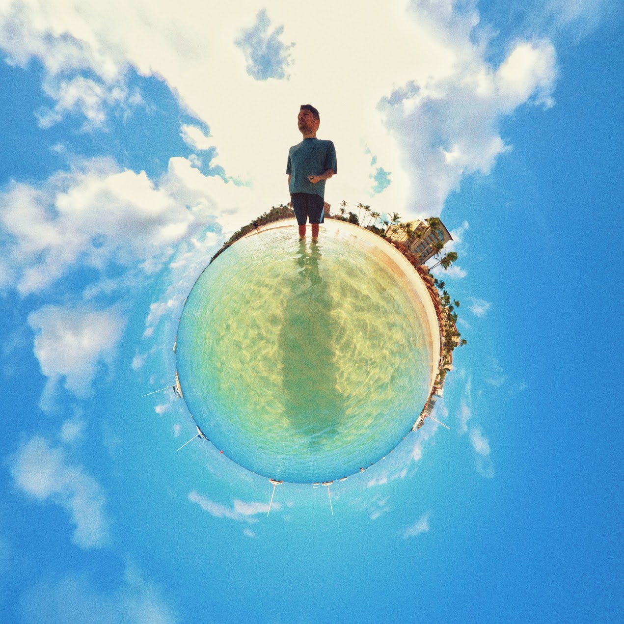 Reza standing on a tropical beach, photographed with a fisheye 'tiny planet' effect showing the shoreline curved into a sphere beneath a blue sky