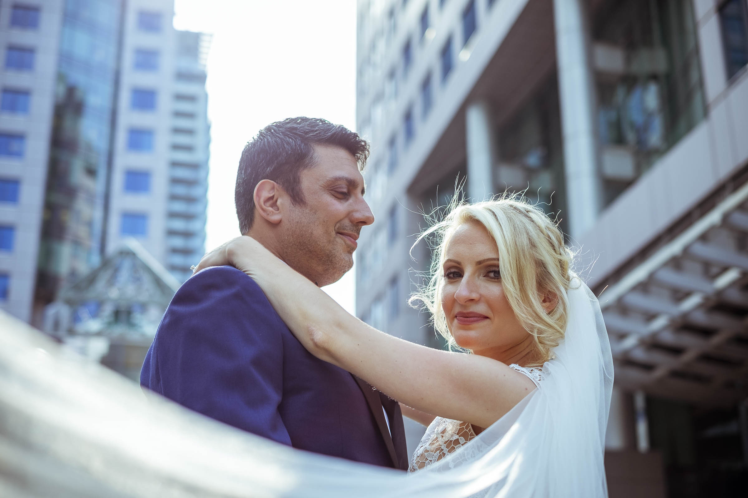 Bride and groom embracing on a rooftop with downtown Toronto skyscrapers in the background, her veil blowing in the wind